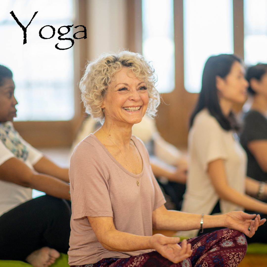 The picture shows an older woman white in the foreground and an African American and Asian woman in the background sitting crossed-legged o the floor with their hands in Ryan mudra in a yoga class.